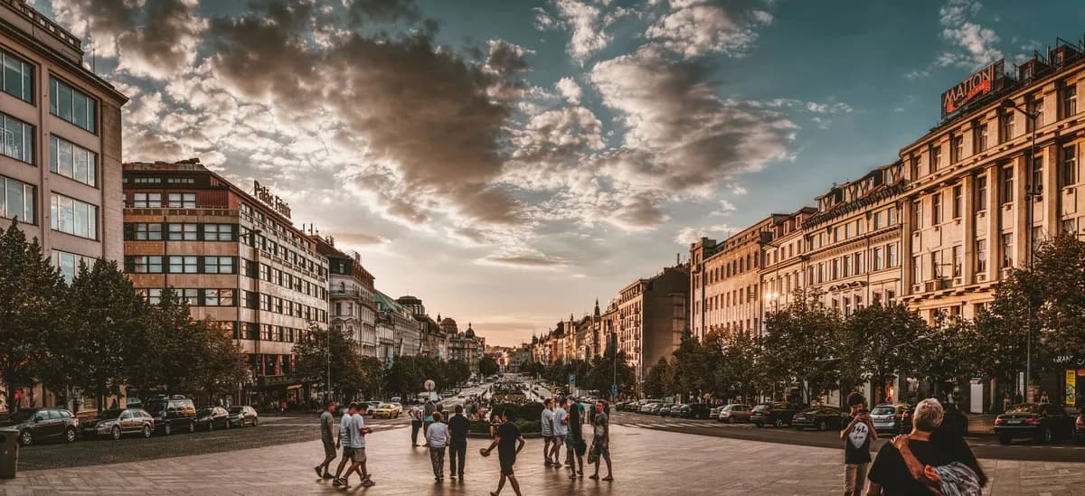 A lively city square at sunset with people walking, historic architecture, and dramatic cloudy sky.